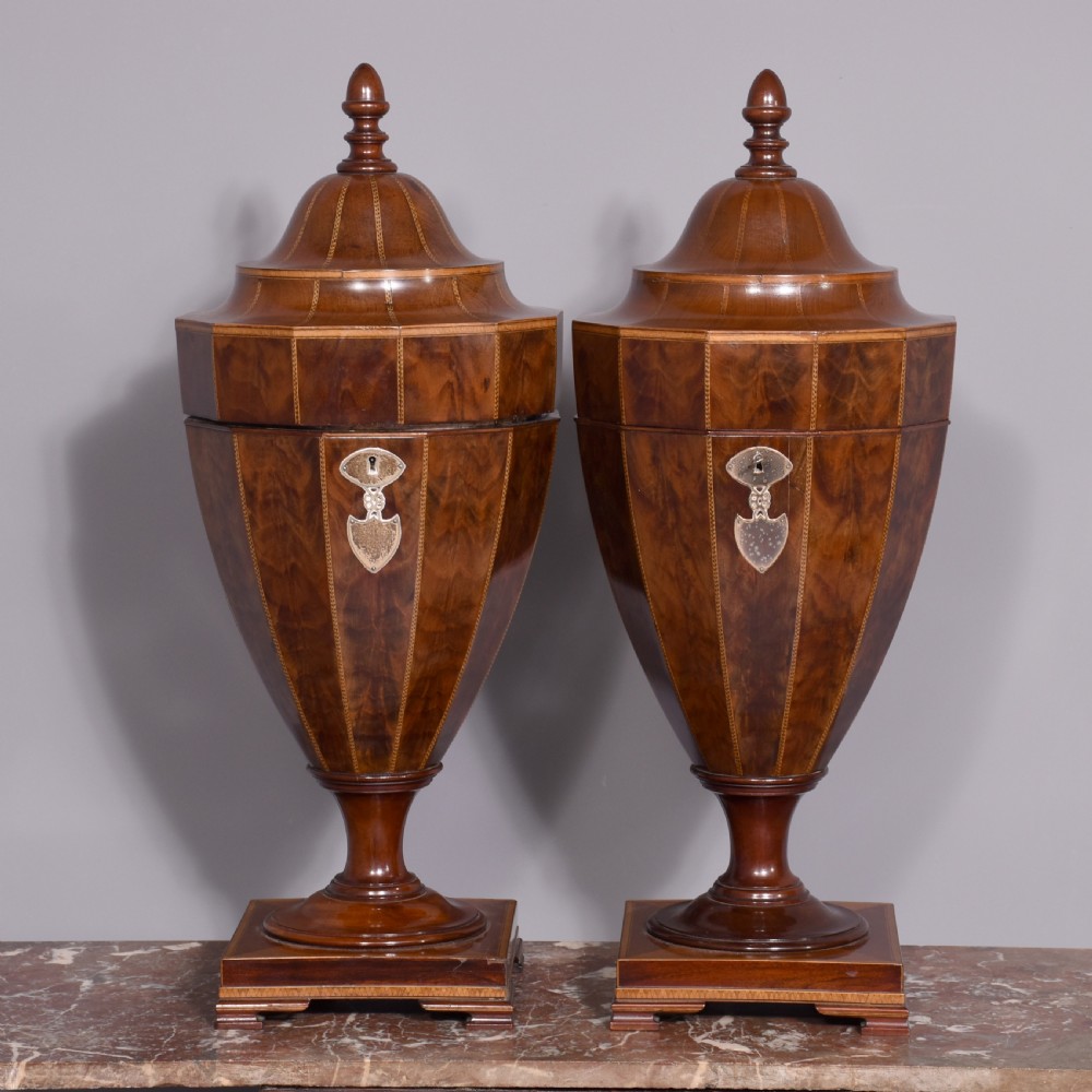 A pair of faceted mahogany cutlery urns featuring geometric stringing inlay, turned finials, and silver keyhole escutcheons on square pedestals.