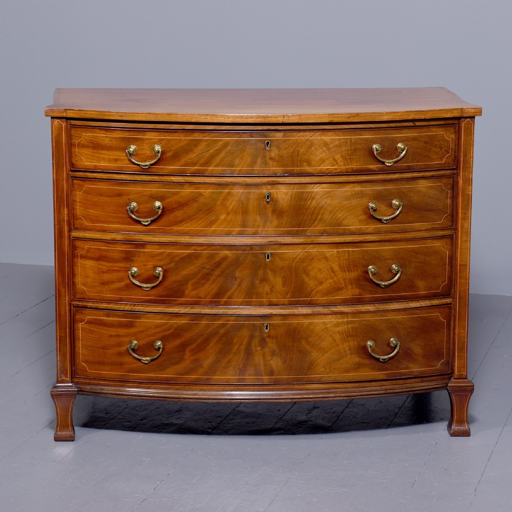 A four-drawer mahogany bow-fronted chest featuring delicate boxwood stringing inlay, brass handles, and splayed bracket feet against a neutral studio backdrop.