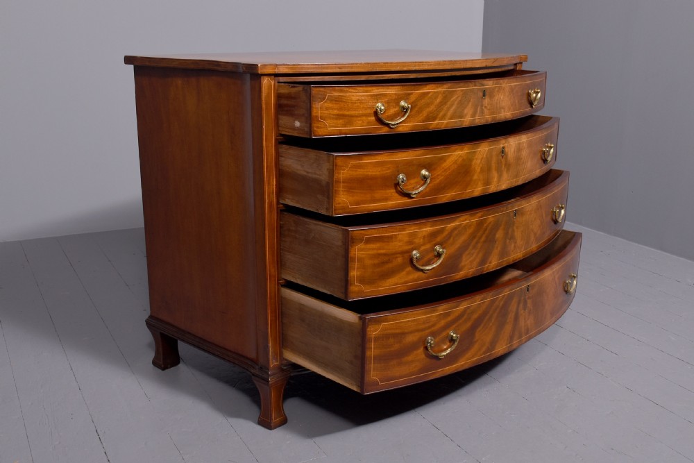 A four-drawer mahogany bow-fronted chest featuring delicate boxwood stringing inlay, brass handles, and splayed bracket feet against a neutral studio backdrop. View V