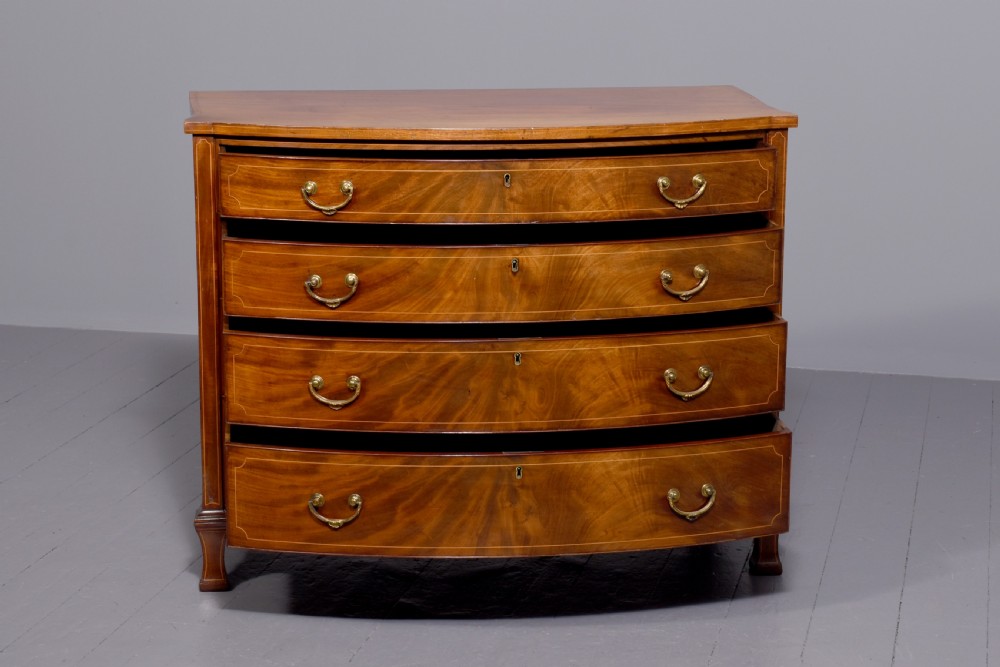 A four-drawer mahogany bow-fronted chest featuring delicate boxwood stringing inlay, brass handles, and splayed bracket feet against a neutral studio backdrop. View VI