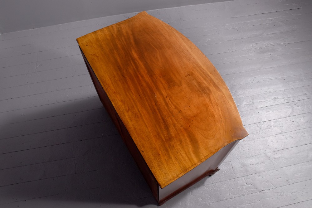 A four-drawer mahogany bow-fronted chest featuring delicate boxwood stringing inlay, brass handles, and splayed bracket feet against a neutral studio backdrop. View VII