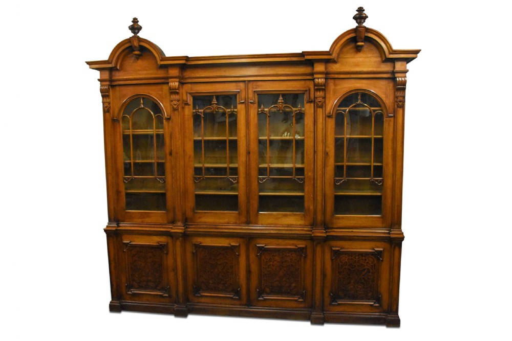 A large Mid-Victorian carved wooden glazed bookcase featuring four upper cabinet doors with arch tracery and four lower panelled cupboard doors topped with decorative finials.
