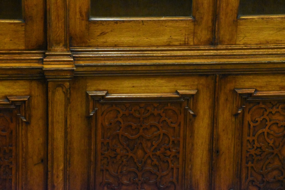 A large Mid-Victorian carved wooden glazed bookcase featuring four upper cabinet doors with arch tracery and four lower panelled cupboard doors topped with decorative finials. View VI
