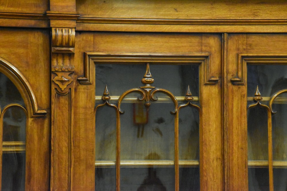 A large Mid-Victorian carved wooden glazed bookcase featuring four upper cabinet doors with arch tracery and four lower panelled cupboard doors topped with decorative finials. View VIII