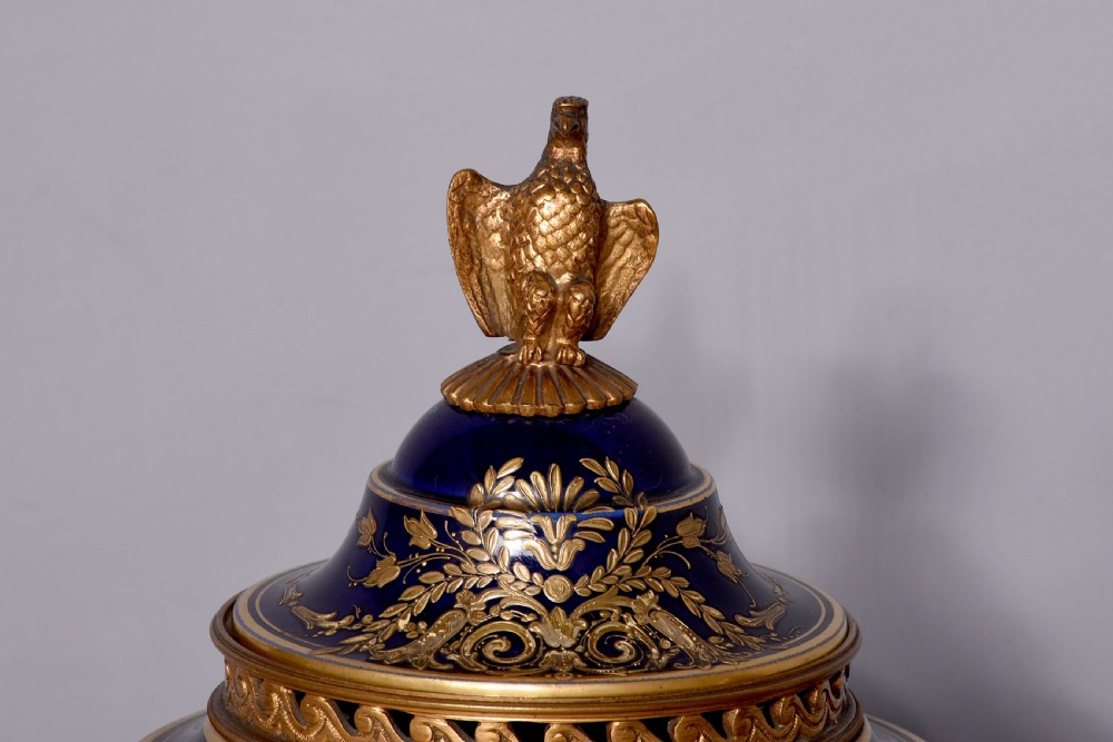 A pair of French cobalt blue porcelain lidded urns with intricate gilding, hand-painted Napoleonic historical scenes, and gilt bronze helmeted bust handles topped with eagle finials. View V