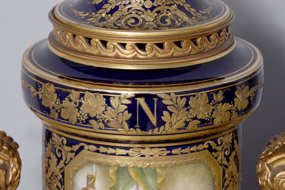 A pair of French cobalt blue porcelain lidded urns with intricate gilding, hand-painted Napoleonic historical scenes, and gilt bronze helmeted bust handles topped with eagle finials. View IX