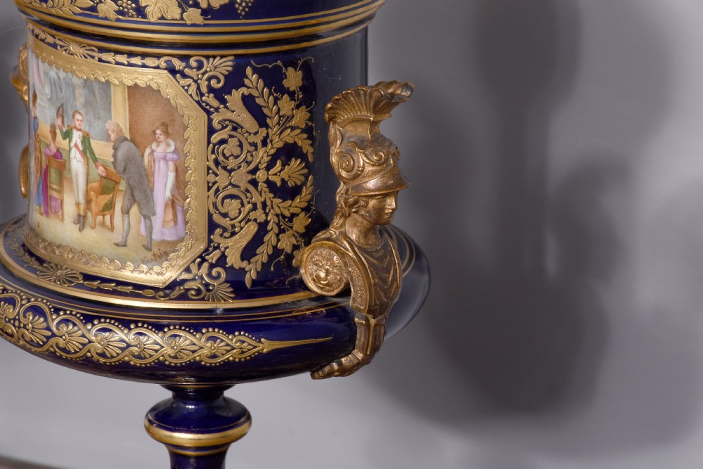 A pair of French cobalt blue porcelain lidded urns with intricate gilding, hand-painted Napoleonic historical scenes, and gilt bronze helmeted bust handles topped with eagle finials. View XI