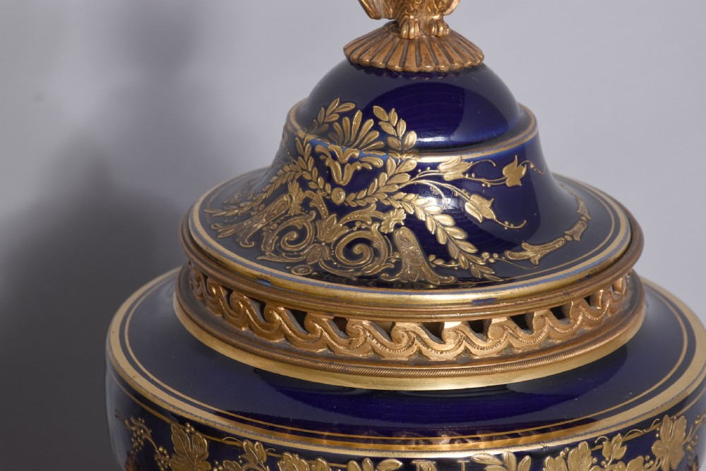 A pair of French cobalt blue porcelain lidded urns with intricate gilding, hand-painted Napoleonic historical scenes, and gilt bronze helmeted bust handles topped with eagle finials. View XVII