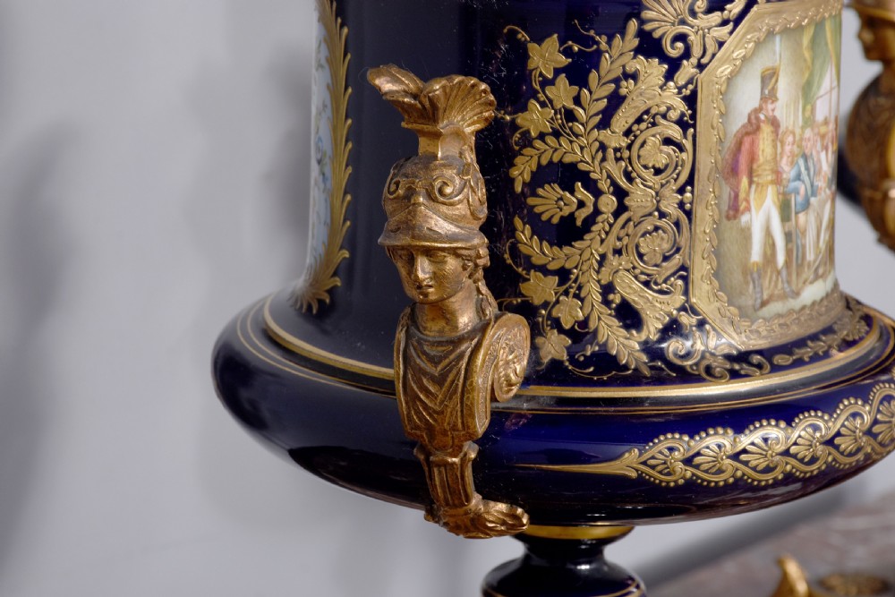 A pair of French cobalt blue porcelain lidded urns with intricate gilding, hand-painted Napoleonic historical scenes, and gilt bronze helmeted bust handles topped with eagle finials. View XIX