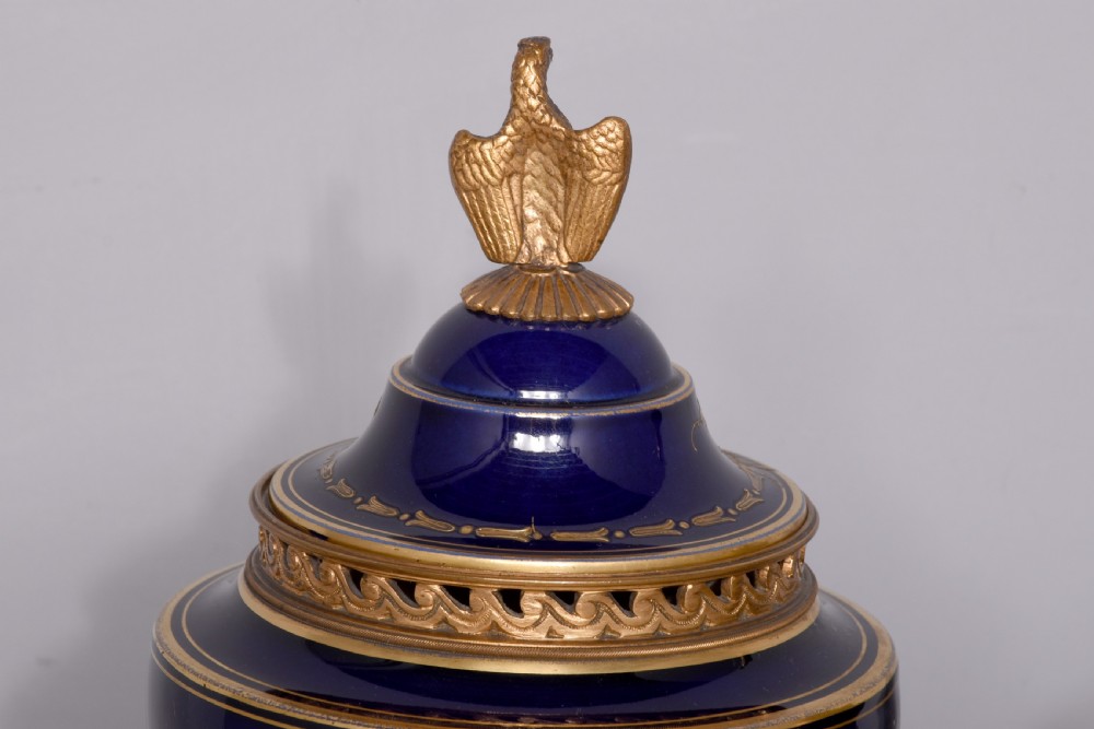 A pair of French cobalt blue porcelain lidded urns with intricate gilding, hand-painted Napoleonic historical scenes, and gilt bronze helmeted bust handles topped with eagle finials. View XXI