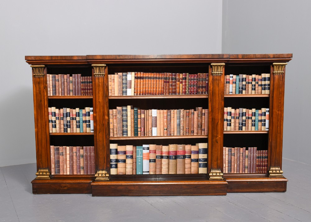 An antique open breakfront bookcase constructed from dark rosewood with gilded carved capitals and filled with leather-bound books.