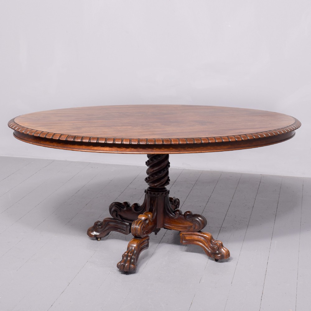 A circular George IV rosewood breakfast table featuring a carved edge, a spiral column pedestal, and a quadruped scrolled base with brass castors.