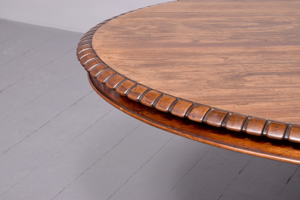 A circular George IV rosewood breakfast table featuring a carved edge, a spiral column pedestal, and a quadruped scrolled base with brass castors. View II