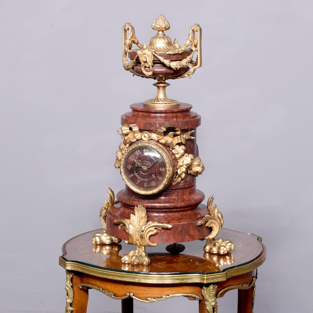A French Neoclassical style mantel clock featuring a cylindrical rouge marble body with gilt bronze floral swags, a top urn finial, and paw feet.