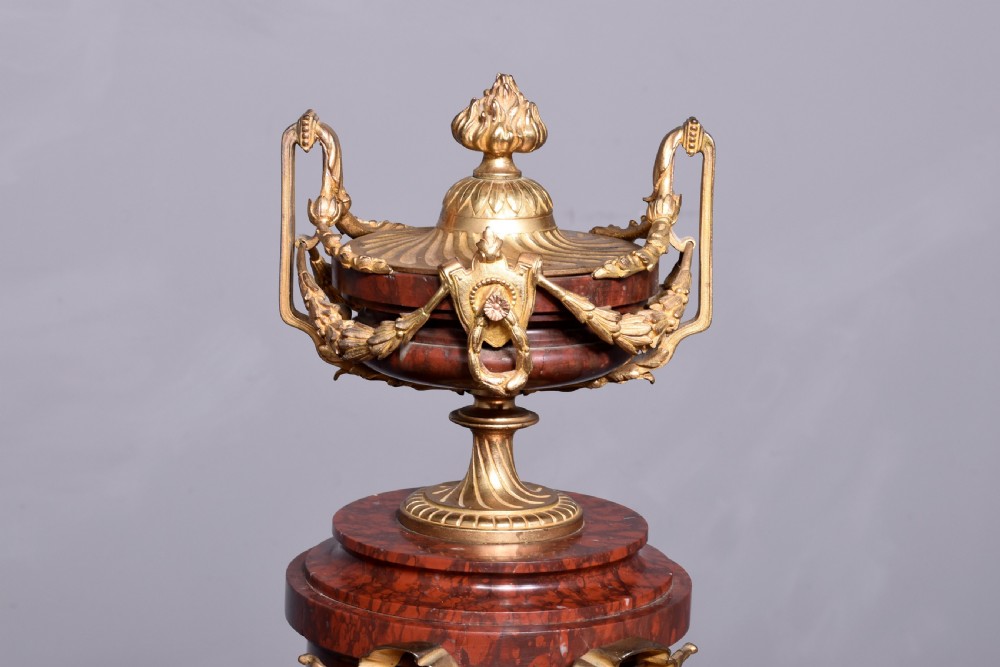 A French Neoclassical style mantel clock featuring a cylindrical rouge marble body with gilt bronze floral swags, a top urn finial, and paw feet. View II