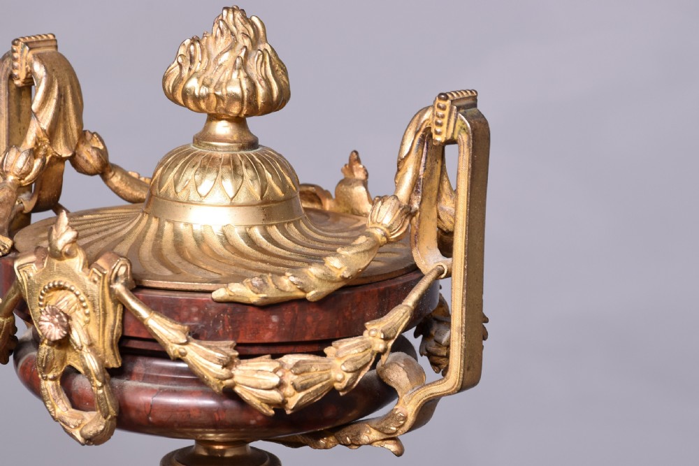 A French Neoclassical style mantel clock featuring a cylindrical rouge marble body with gilt bronze floral swags, a top urn finial, and paw feet. View V