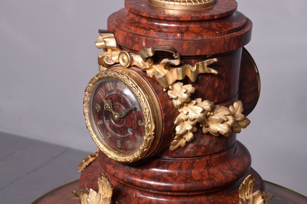 A French Neoclassical style mantel clock featuring a cylindrical rouge marble body with gilt bronze floral swags, a top urn finial, and paw feet. View VI