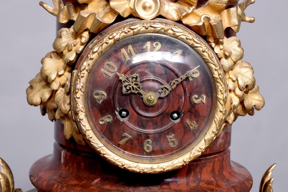 A French Neoclassical style mantel clock featuring a cylindrical rouge marble body with gilt bronze floral swags, a top urn finial, and paw feet. View X