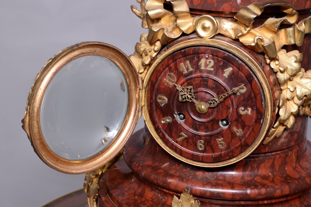 A French Neoclassical style mantel clock featuring a cylindrical rouge marble body with gilt bronze floral swags, a top urn finial, and paw feet. View XI