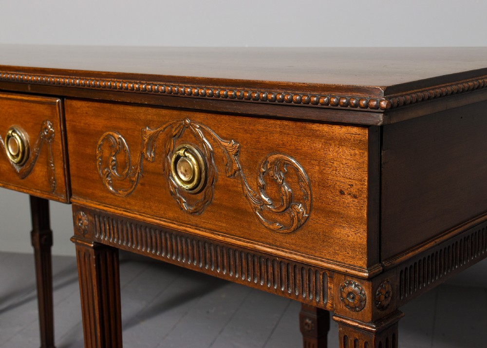 A long Georgian mahogany console table featuring a carved frieze with urn motifs and swags, brass ring handles, and fluted legs. View II