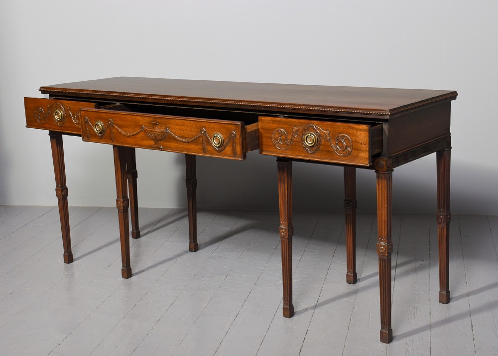A long Georgian mahogany console table featuring a carved frieze with urn motifs and swags, brass ring handles, and fluted legs. View IV