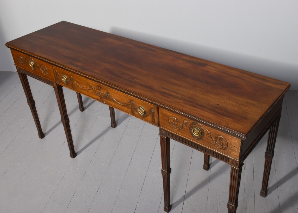 A long Georgian mahogany console table featuring a carved frieze with urn motifs and swags, brass ring handles, and fluted legs. View V