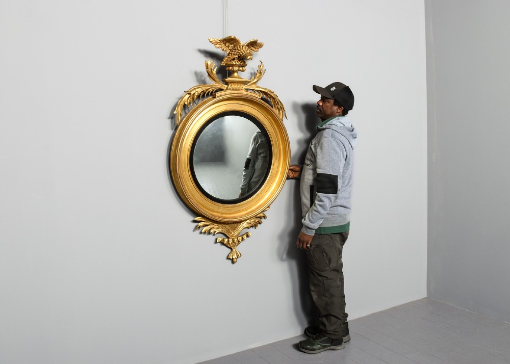 A circular Regency period giltwood convex mirror framed with an ebonised inner slip ring, topped by a carved eagle surmount perched on a pedestal with acanthus leaf accents. View III