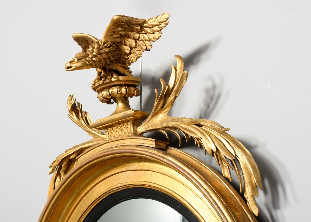 A circular Regency period giltwood convex mirror framed with an ebonised inner slip ring, topped by a carved eagle surmount perched on a pedestal with acanthus leaf accents. View V