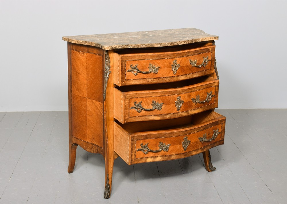 An antique French Louis XV style serpentine-fronted commode featuring three drawers with geometric wood inlay, cast brass handles, and a fitted beige marble top. View II