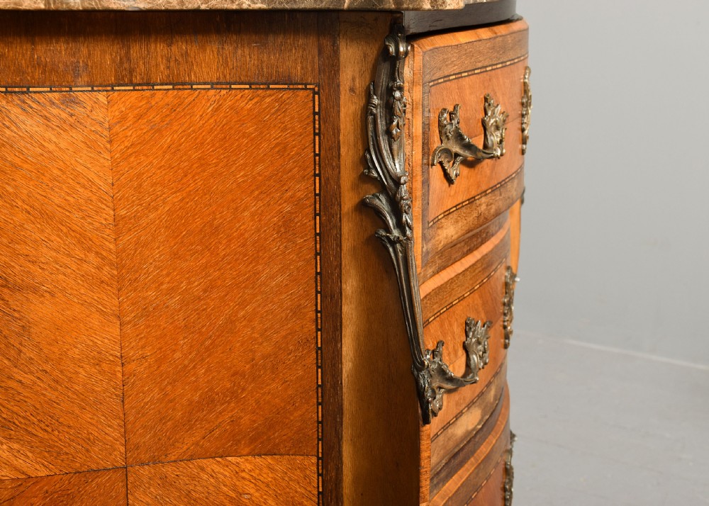 An antique French Louis XV style serpentine-fronted commode featuring three drawers with geometric wood inlay, cast brass handles, and a fitted beige marble top. View IV