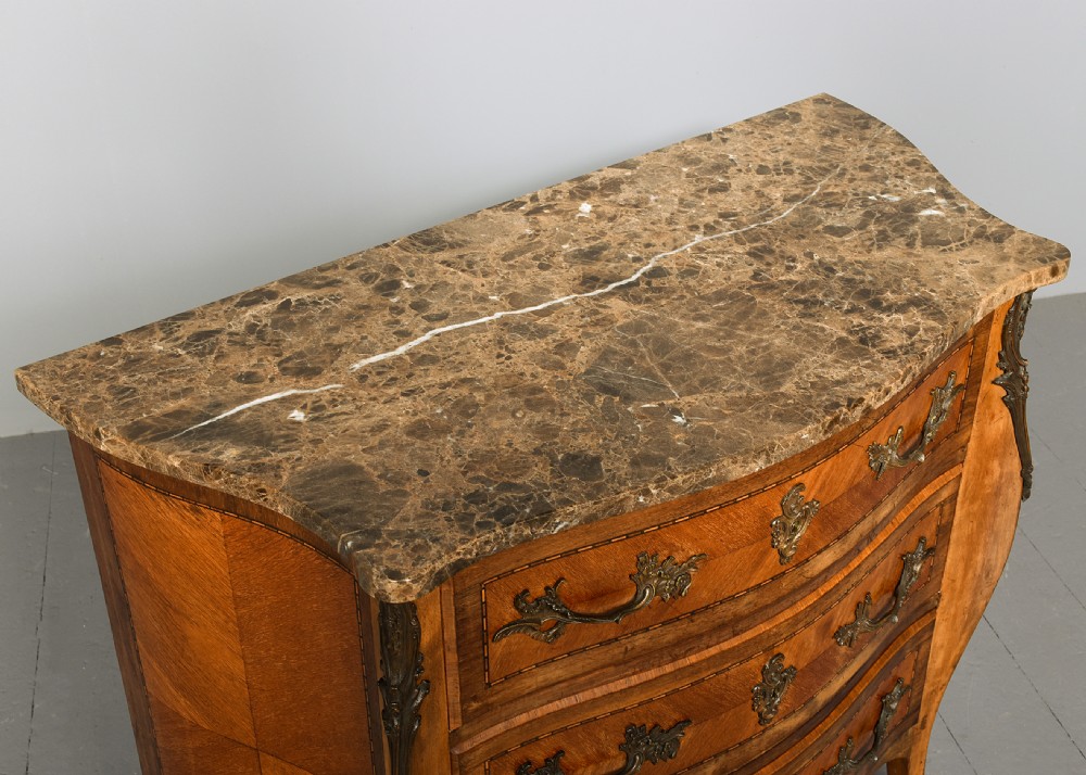 An antique French Louis XV style serpentine-fronted commode featuring three drawers with geometric wood inlay, cast brass handles, and a fitted beige marble top. View IX