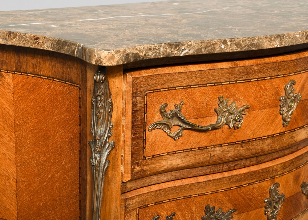 An antique French Louis XV style serpentine-fronted commode featuring three drawers with geometric wood inlay, cast brass handles, and a fitted beige marble top. View X