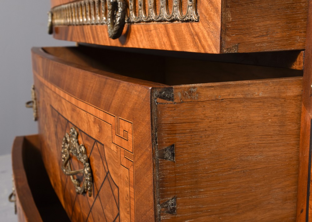 A French marquetry-inlaid walnut commode featuring three drawers, ornate brass hardware, and a brown veined marble top. View III