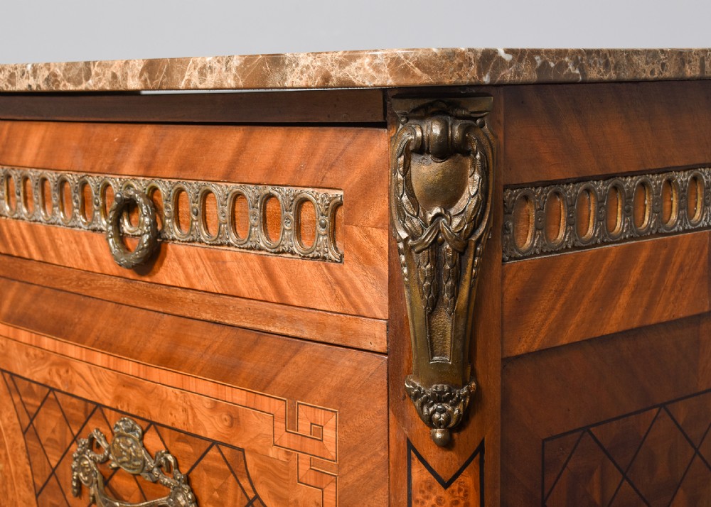 A French marquetry-inlaid walnut commode featuring three drawers, ornate brass hardware, and a brown veined marble top. View IV