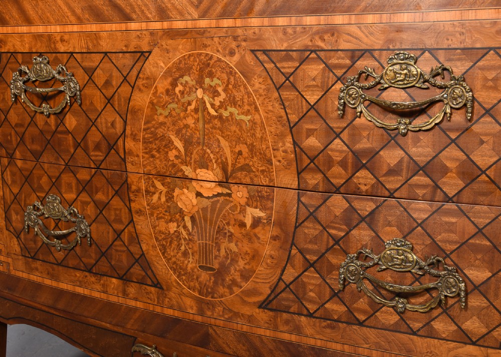 A French marquetry-inlaid walnut commode featuring three drawers, ornate brass hardware, and a brown veined marble top. View V