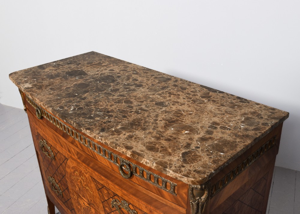 A French marquetry-inlaid walnut commode featuring three drawers, ornate brass hardware, and a brown veined marble top. View IX