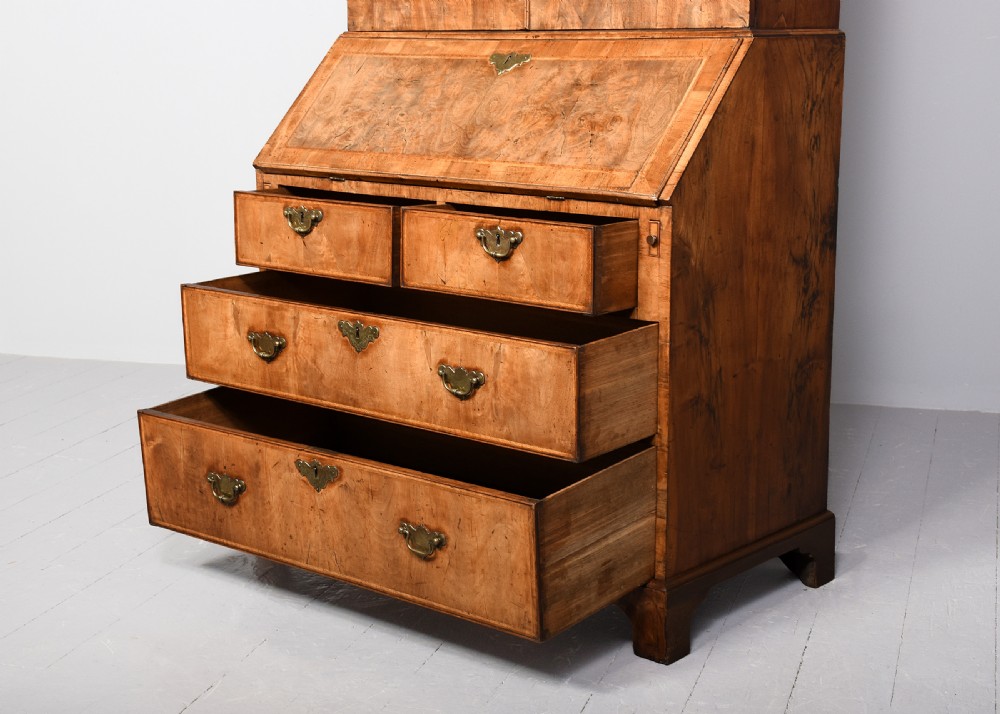 A George II walnut bureau bookcase featuring a broken arch pediment with finial, glazed upper doors, a fall-front desk, and lower drawers on bracket feet. View X