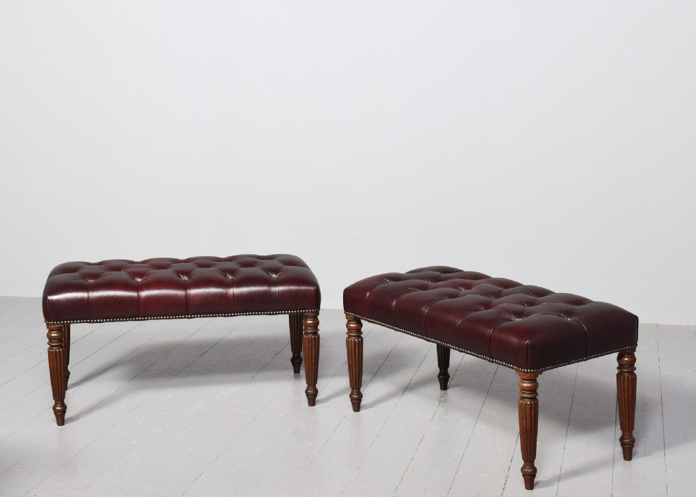 A pair of rectangular mahogany long stools upholstered in deep-buttoned burgundy leather with brass stud borders and fluted turned legs.