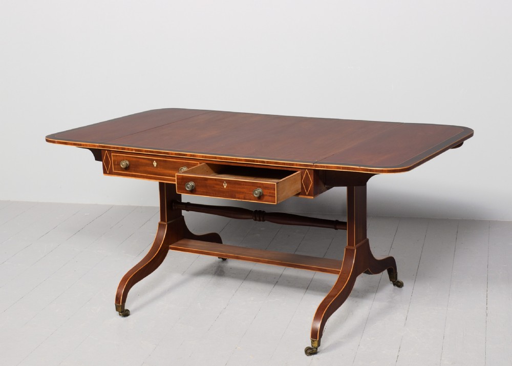 A Regency mahogany sofa table featuring drop flaps, geometric string inlay, twin frieze drawers, and splayed legs with brass castors. View III