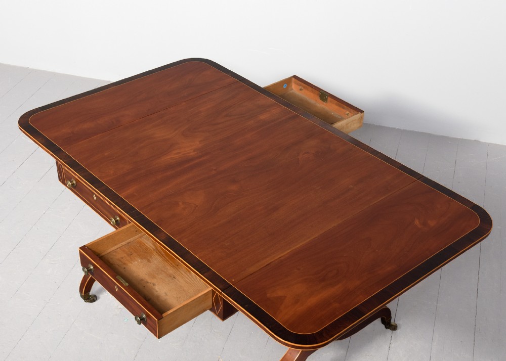 A Regency mahogany sofa table featuring drop flaps, geometric string inlay, twin frieze drawers, and splayed legs with brass castors. View IV