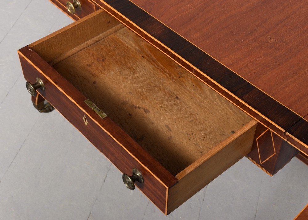 A Regency mahogany sofa table featuring drop flaps, geometric string inlay, twin frieze drawers, and splayed legs with brass castors. View V