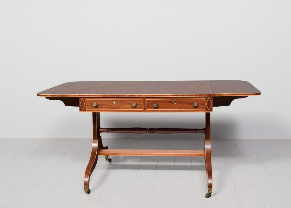 A Regency mahogany sofa table featuring drop flaps, geometric string inlay, twin frieze drawers, and splayed legs with brass castors. View X