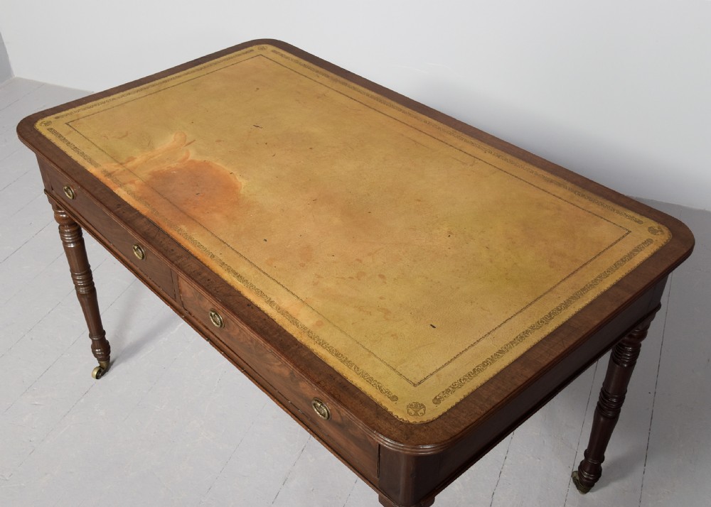 A George III mahogany library table featuring a tan leather writing surface, two frieze drawers with brass ring pulls, turned legs, and brass castors. View II