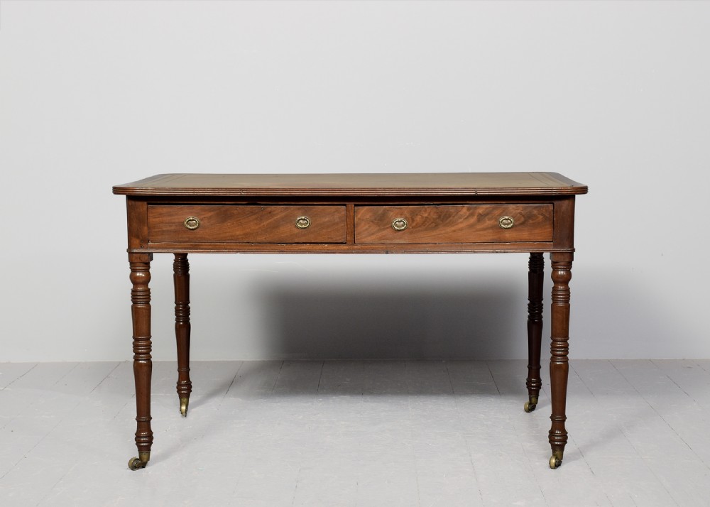 A George III mahogany library table featuring a tan leather writing surface, two frieze drawers with brass ring pulls, turned legs, and brass castors. View V