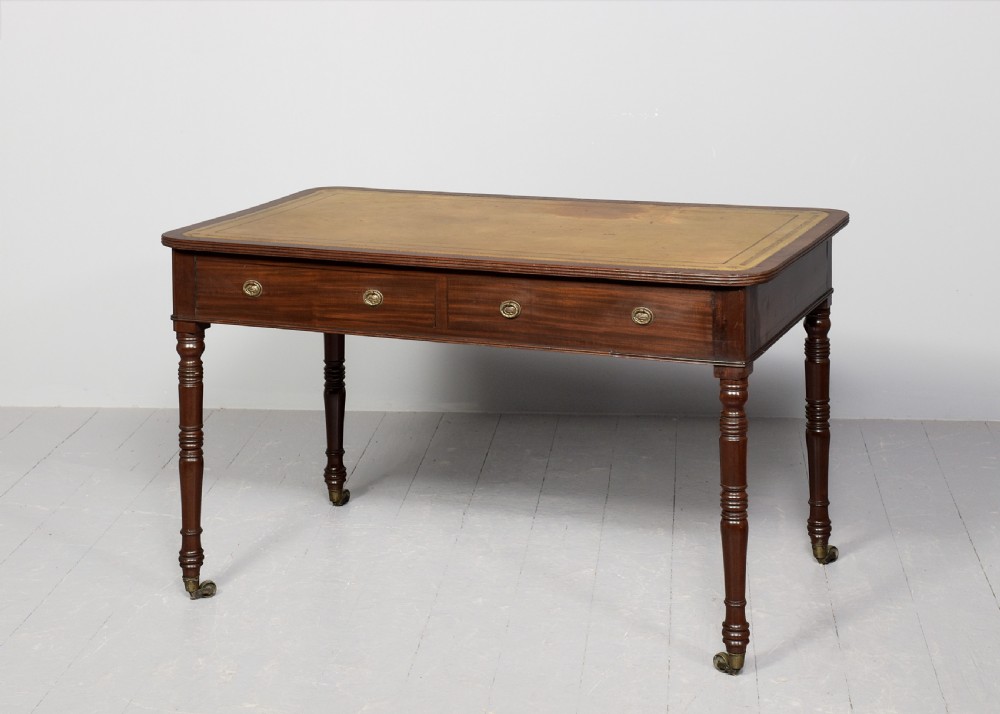 A George III mahogany library table featuring a tan leather writing surface, two frieze drawers with brass ring pulls, turned legs, and brass castors. View VI