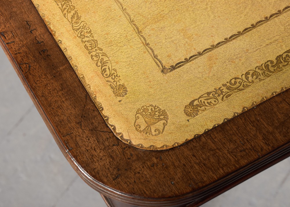 A George III mahogany library table featuring a tan leather writing surface, two frieze drawers with brass ring pulls, turned legs, and brass castors. View X
