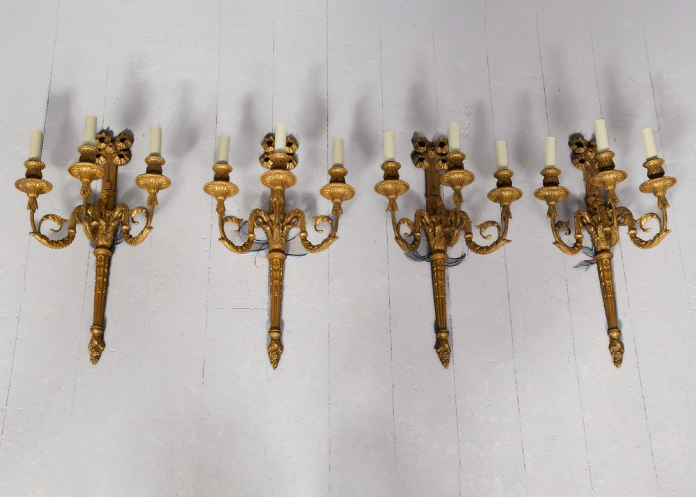 A set of four gilded three-arm wall sconces featuring neoclassical ribbon bow tops and scrolling leaf motifs. View VIII