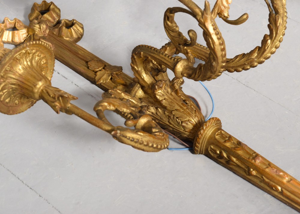 A set of four gilded three-arm wall sconces featuring neoclassical ribbon bow tops and scrolling leaf motifs. View X