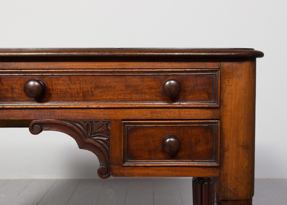 A George IV mahogany writing table featuring four drawers with wooden turned handles, carved corner spandrels, and reeded turned legs. View IV