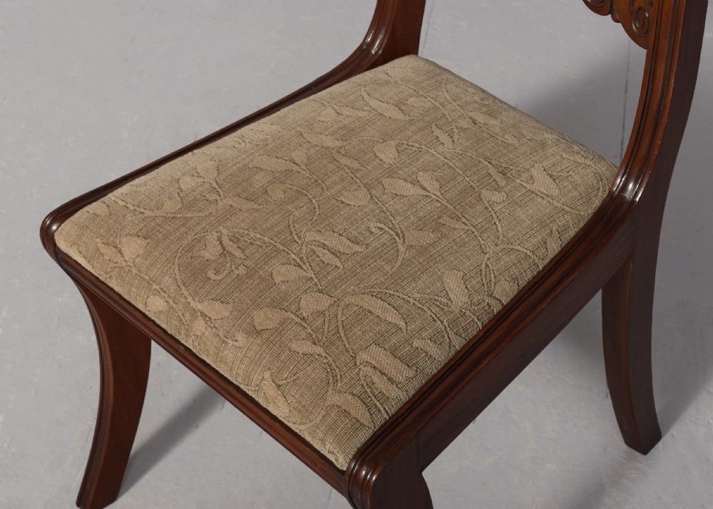 A set of ten Scottish carved mahogany dining chairs featuring cresting bar backs, carved splats, neutral fabric seats, and curved sabre front legs. View X